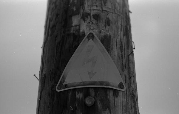 Black and white photography of a triangle sign with a lightning in it on an wooden electrical pole.