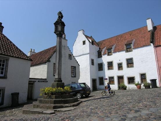 The mercat cross in Culross. The image shows a square in a village paved with large cobbles and sloping from right down to left. The mercat cross dominates the left centre of the frame and there’s a child on a bicycle to its right. In the background and on the left are white painted old buildings with red pantiled roofs. The sky is blue.