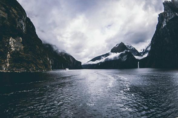 L'eau paisible frémis à la surface. De chaque côté, les montagnes et roches néo-zélandaises entourent le voyageur. Le sommet comme le ciel est empli de nuages blancs.