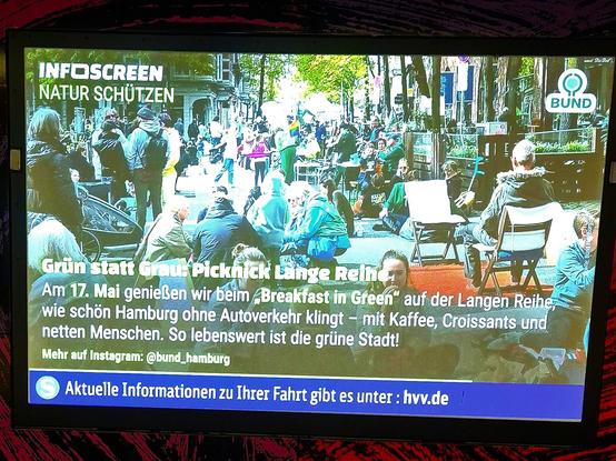 Infoscreen-Anzeige am S-Bahnsteig, zu sehen ist der Screen und ein bisschen rot-schwarz gemusterte Haltestellen-Wand. auf dem Screen befindet sich ein Bild, auf denen viele Menschen bei einem Picknick auf der Straße sitzen, stehen und herumlaufen. Zwei helle Klappstühle stehen auf einem roten Stück roten Teppich. Die Menschen sind größtenteils in Grüntönen gekleidet. Auf dem Bild schwebt in weißer Schrift:
Grün statt Grau: Picknick Lange Reihe
Am 17. Mai genießen wir beim "Breakfast in Green" auf der Langen Reihe,
wie schön Hamburg ohne Autoverkehr klingt - mit Kaffee, Croissants und
netten Menschen. So lebenswert ist die grüne Stadt!
Mehr auf Instagram: @bund_hamburg
Oben rechts in klein das Logo vom BUND (Bund für Umwelt und Naturschutz). Und links steht das Wort "Infoscreen" und darunter "Umwelt schützen".