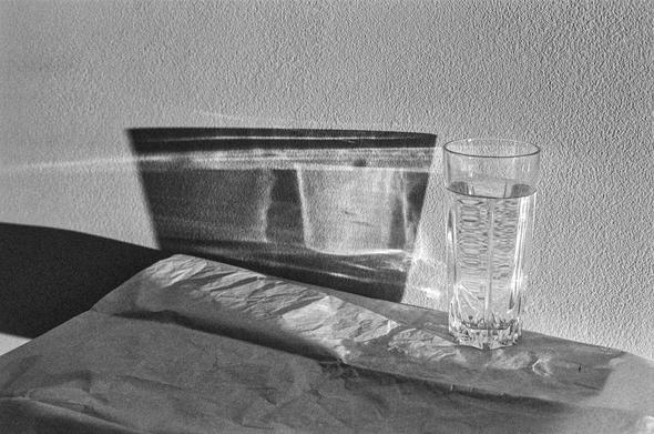 The black-and-white image shows a clear glass of water placed on a crumpled piece of paper. The surface is smooth and flat, and the background is a textured wall. Light passing through the glass creates intricate reflections and refractions on the wall, forming abstract shapes and patterns. The lighting suggests natural sunlight coming from the right side, casting shadows and creating a striking contrast that adds a sense of depth and calmness to the composition.