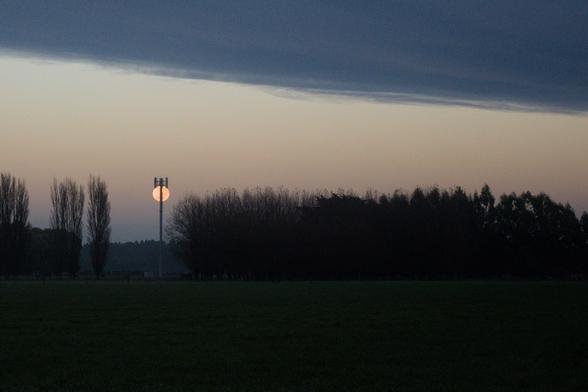 A photo of farmland between Christchurch and Lincoln, New Zealand, with the near-full moon setting behind a cellphone tower.