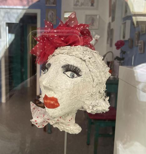 Sculpture of a woman's head made of white material. The lips are painted red, eyes and eyelashes are highlighted in color. The sculpture is wearing a red dotted scarf around her neck and a red hairband on her head.