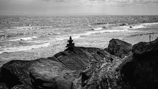 A black and white photograph of a rocky shelf with a pebble tower looking out to sea across to distant land.