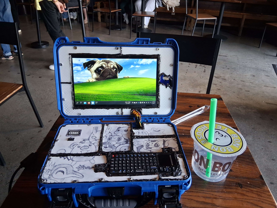 Cyberdeck on a desk, consisting of a blue pelican case with a screen displaying a pug haphazardly glued into place, and on the bottom half a mini keyboard also glued on with the gaps filled in by white panelling with hand drawings on it