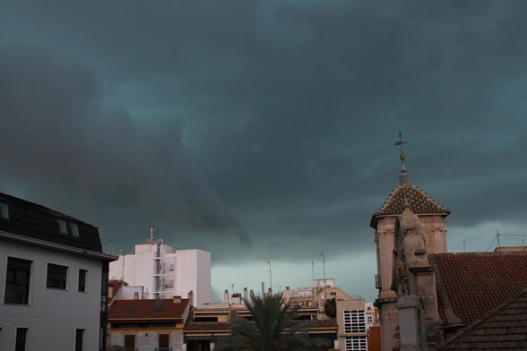 Un paisaje urbano situado en la ciudad de Murcia. Se aprecian edificios y la torre campanario de una iglesia. El cielo, azulado, está cubierto de nubes oscuras que anuncian una inminente tormenta, y a lo lejos se ve el cielo un poco más despejado y el sol ilumina un edificio blanco mientras el resto está en penumbra.