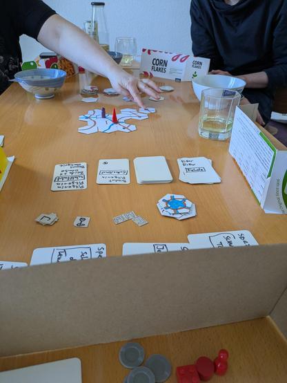 Aufnahme eines Spieltisches mit einigen sechseckigen Plättchen und ausliegenden Karten und Markern. Alles sieht selbstgebastelt aus. Auf den Sechsecken stehen zwei Spielfiguren. Eine Hand ist im Bild.
