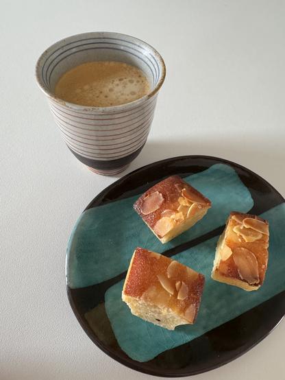 Trois morceaux d’un financier aux amandes dans un joli petit pat japonais en céramique turquoise et couleur café, à côté d’une tasse à café allongé grisé aux rayures rouges dehors et bleues dedans