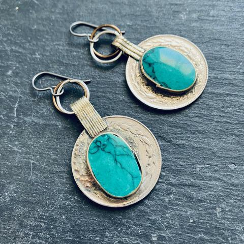 A photo of a pair of coin-shaped, silver toned dangle earrings on a slate grey background. The coin earrings are made of mixed metals with turquoise-like blue oval stones affixed to them.