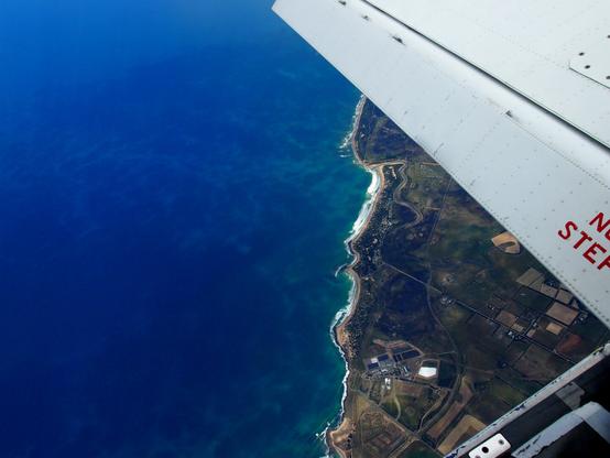 Image of a coastline taken from the open door of a skydiving aircraft flying at 15,000 feet. The images foreground shows a small portion of the flap side of the left wing with NO STEP printed in red and the edge of the door sill. Far below is a crystal blue ocean, a portion of coastline and green fields next to the coast.