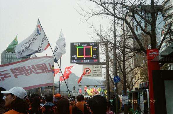 광화문 일대에 도착한 민주노총 총파업 행진 대오. 여러 깃발과 현수막이 나부끼는 가운데 저 멀리 북악산과 이순신 장군 동상이 보인다. 전광판에 광화문 일대가 정체중이라는 그래픽이 떠있다.