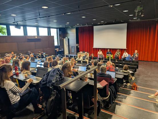 Foule assise dans un amphi devant un panel de 5 personnes assises sur des fauteuils sur scène