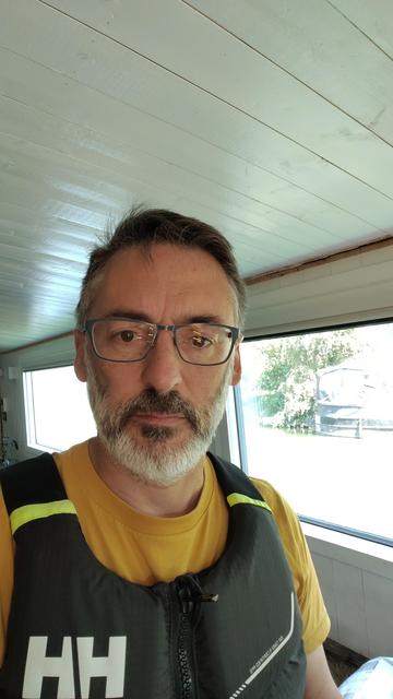 A white man in his 50's with dark hair, glasses, an orange t-shirt and a Helly Hansen bouyancy aid stood in a boat, in the background through a window you can see another canal boat.