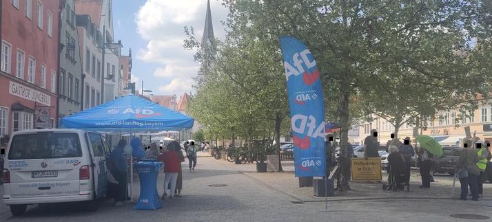 In einer Innenstadt ist auf der linken Seite ein Infostand der AFD: ein weißer Bus, ein blauer Sonnenschirm mit Parteilogo, mehrere Personen stehen darunter, ein paar Meter weiter weht eine blaue Fahne mit Parteilogo
Auf der anderen Seite ist eine Gegenkundgebung: ca. zehn Personen mit Transparenten mit Aufschriften wie "Lasst euch nicht betören von ihren schönen Worten"
Im Hintergrund stehen mehrere große, prächtig blühende Bäume
Außerdem sind ein hellblauer Himmel mit weißen Wolken und die Spitze eines Kirchturms zu erkennen
