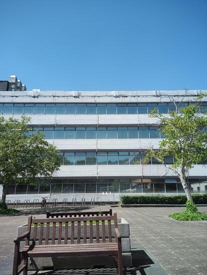 View of nondescript 60s concrete building with two trees and bench in foreground