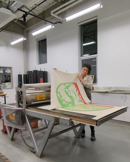 Artist Jenie Gao in the print shop, pulling a large scale woodblock print (in jade green and red) on the press. The image is of a homing pigeon and resembles carved jade.

The print is roughly 4x3 feet and on muslin canvas.

Jenie is of Taiwanese and Chinese descent and has short black hair with a side fade, and is wearing an inky lab coat.