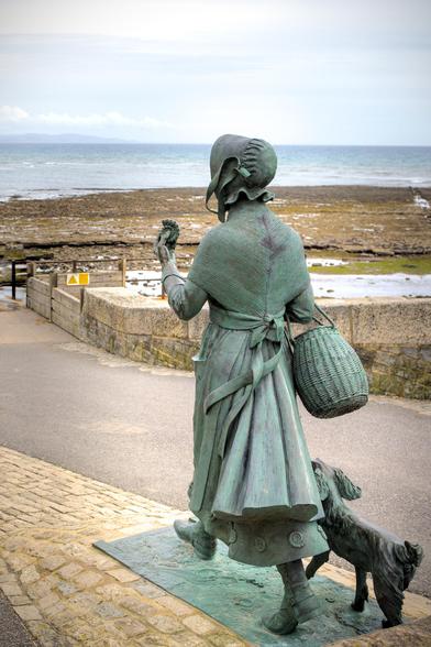 A statue of a woman, seen from behind walking along a path above a beach. From behind, her head and face are not visible - she is wearing a bonnet. The statue includes a dog running at her feet, She is holding up a fossil of an ammonite in her hand.