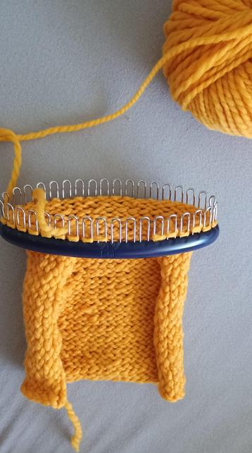 Photo of a yellow piece of knitting on a blue oval loom with metal pegs. At the top right a part of the yarn ball is visible.