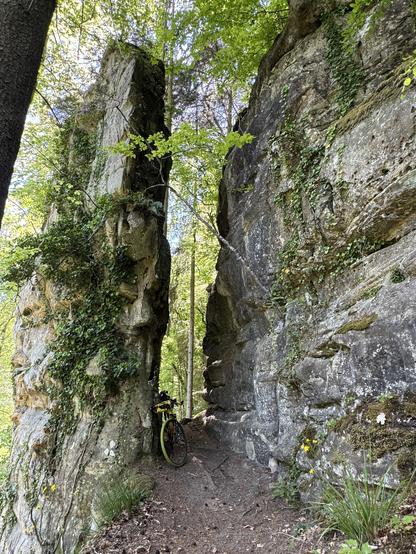 My gravel bike is between two huge and tall rocks, leaning against the one on the left. There is quite a narrow passage between the rocks. Greenery is growing on and around them.