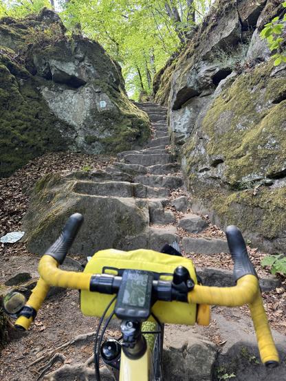 In the foreground my bike’s yellow drop handlebars, looking at steep irregular steps going up between rocks. Yes, my bike is looking. Greenery is visible at the top of the steps.