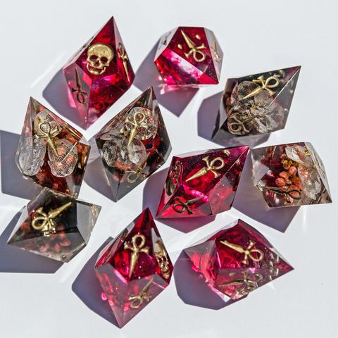 Hier ist das 10-teilige Vampire Würfelset auf weißem Untergrund zu sehen. Die Hunger Würfel sind in Blutrot mit goldenen Symbolen gehalten und zeigen neben dem Ankh-Symbol ein Vampirschädel Symbol. Die normalen Würfel haben Blüten im inneren und sind in einem transparenten schwarz gehalten.