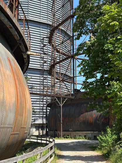 Ein Weg führt in einer Kurve nach links zwischen zwei großen, rostigen, runden, bauchigen Industriebauten (Teleskopgasbehälter) hindurch, rechts im Vordergrund noch eingerahmt durch einen Baum. Im Hintergrund ragt ein noch viel größeres Gebäude in die Höhe, grau und nicht verrostet (Scheibengasbehälter)