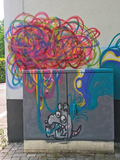 A grey electrical box is covered in colourful graffiti. Abstract shapes in red, pink, yellow, and blue swirl across the box, resembling spaghetti. A cartoon rat character is painted at the bottom, appearing to eat the colourful swirls. The rat is grey with exaggerated features.
