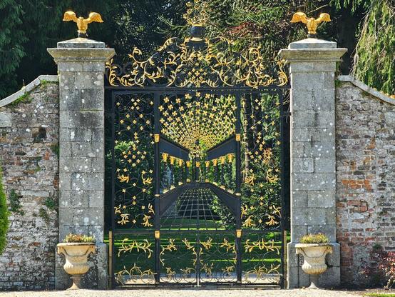 An ornate black wrought iron gate with gilded floral and scroll details stands open, flanked by weathered stone pillars each topped with a golden eagle. Beyond the gate, a tree-lined path leads into a lush green garden. Brick and stone walls enclose the scene.