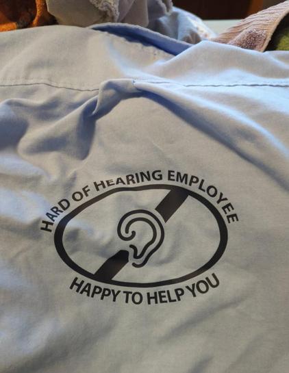 A folded light blue shirt with a black logo. The logo shows an oval containing an ear symbol with a line through it, above the words "Happy to Help You" and below the words "Hard of Hearing Employee."