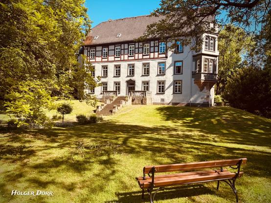 Blick über eine Parkbank hinweg auf das Schloss Großen-Buseck, eingerahmt von alten Bäumen – ein ruhiger Ort, der zum Verweilen und Staunen einlädt.