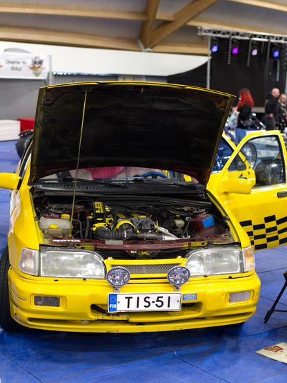 A 1990 Ford Sierra at a car show, with hood open, showcasing the engine. The car is yellow, with checkered-flag decal on doors. Finnish license plate "TIS-51." Background features event lighting and attendees.