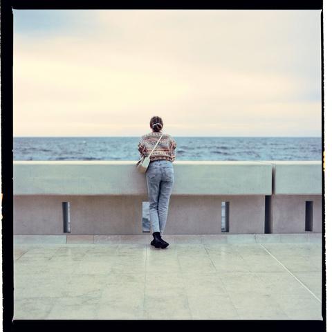 A person sits on a ledge overlooking an ocean with waves crashing against rocks, captured in a photograph taken during daylight.