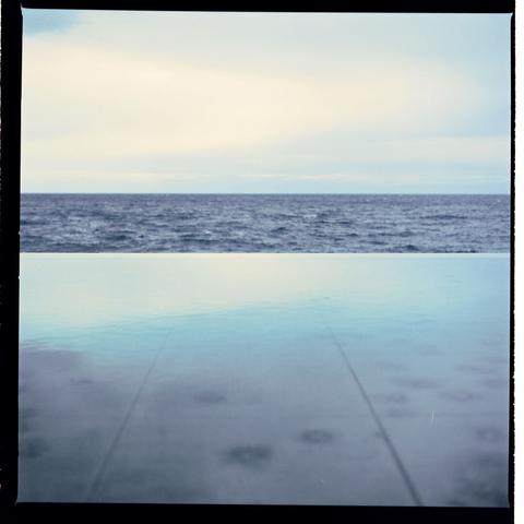 The photo depicts a quiet scene with no people, an overcast sky, and a calm ocean reflected on its surface. A concrete structure resembling a swimming pool can be seen in the foreground.