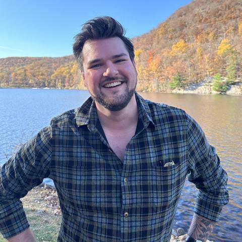 Head-and-shoulders three-quarters view color photo of author Sam Rebelein, a rosy-cheeked man with dark hair worn short and brushed back from his forehead, plus a beard and mustache. He stands in front of a body of water with a hilly autumn landscape on the opposite shore. He wears a blue-and-green plaid shirt and grins into the camera.