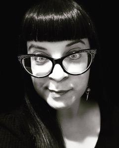 Black-and-white closeup photo portrait of Angela Sylvaine, a fair-skinned woman with pale eyes and straight, dark hair worn long and with bangs. She wears a dark V-neck, long earrings, and large, ornate, cat-eye glasses. She quirks her sharply peaked eyebrows and smiles mischievously into the camera.