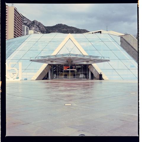 This is an image of a modern architectural structure. The building has a distinctive shape with a curved glass facade and a prominent triangular overhang. In the foreground, there is a wet sidewalk, suggesting recent rain, which adds to the overall moody atmosphere of the photo. The sky is overcast with no visible sunlight, contributing to the subdued lighting in the scene.