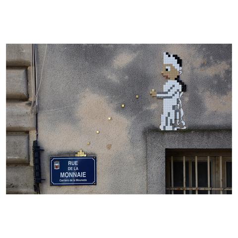 Image of the outer wall of a building next to Palais des Papes in Avignon. The gray wall has a rough texture. In the lower left, a sign reads "Rue de la Monnaie - Carriero de la Mounedo". In the top right, a figure made from small tiles "stands" on top of a window frame. The figure throws coins which have formed a small pile on top of the sign.