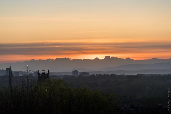 A tranquil sunset over Edinburgh, Scotland, with silhouetted city buildings and church spires in the foreground, lush green trees, and the iconic Forth Bridge visible in the distance beneath a dramatic sky of layered orange, yellow, and grey clouds.