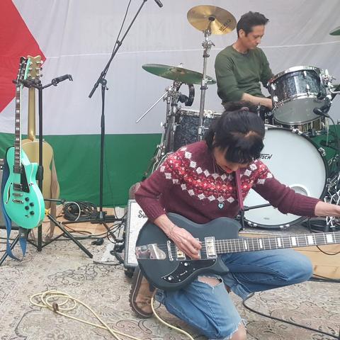 Photograph of Kristin tuning a guitar in the foreground, in the background Fred at the drums. As backdrop a large Palestinian flag. Outside the frame to the left Pete Harvey on cello, and to the right Dylan Hersh Going. Real photographer: me.