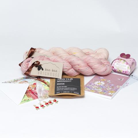 Picture shows two skeins of yarn, one behind the other, in shades of pink with red, yellow and blue speckles. In front of the yarn, from left to right is a card, five rose stitche markers, midnight rose tea, a notebook with flowers on the cover and cherry blossom soap in a gift box.