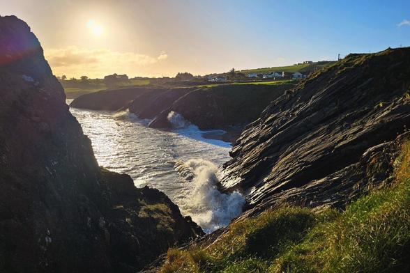 A wide shot of a rugged Irish coastline at sunset, featuring waves crashing against dark, rocky cliffs. The sun is setting over the water, casting a golden glow on the scene.