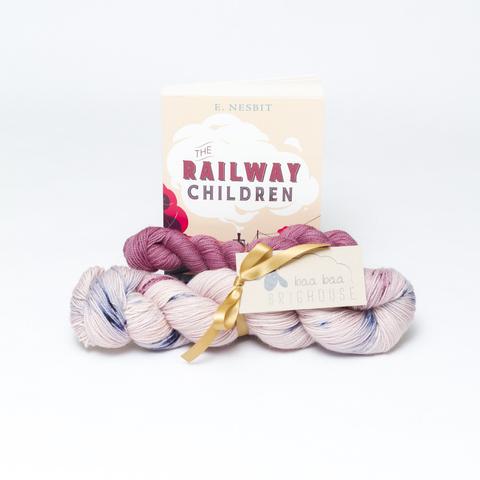 Picture shows a sock set of yarn consisting of main skein which is pink with blue and red speckles and a dark pink mini skein. They are tied together with gold ribbon. Behind the yarn stood on its end is a copy of The Railway Children.