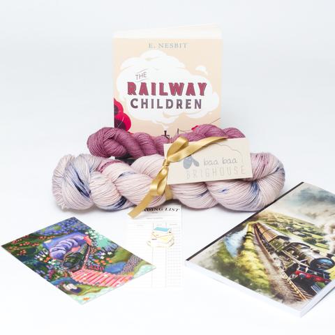 Picture shows a sock set of yarn consisting of main skein which is pink with blue and red speckles and a dark pink mini skein. They are tied together with gold ribbon. Behind the yarn stood on its end is a copy of The Railway Children. In front of the yarn, from left to right is a postcard, a pin badge and a notebook.