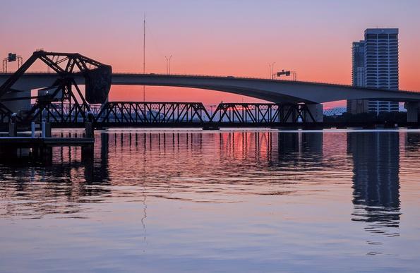 Colorful sunrise glow over the downtown skyline and bridges over the Saint Johns River, casting reflections upon the calm water below.