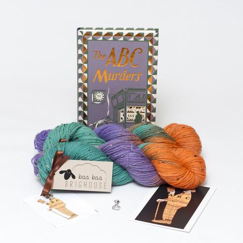 Picture shows to skeins of yarn, one in front of the other, in shades of green, purple and orange. Behind the yarn, stood on its end is a hardback copy of Agatha Christie's The ABC Murders. In front of the yarn, from left to right is a sticker, a letter stitch marker and a postcard.