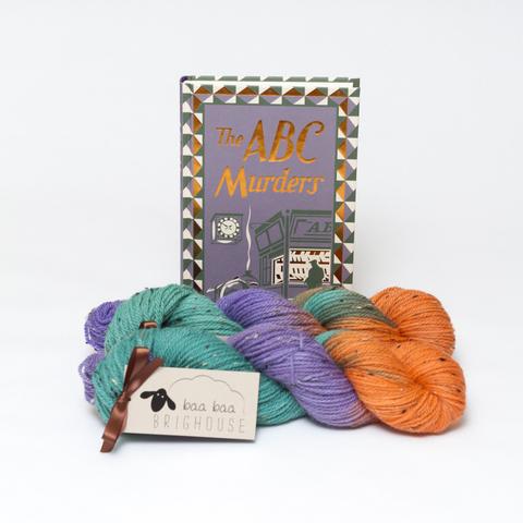 Picture shows to skeins of yarn, one in front of the other, in shades of green, purple and orange. Behind the yarn, stood on its end is a hardback copy of Agatha Christie's The ABC Murders.