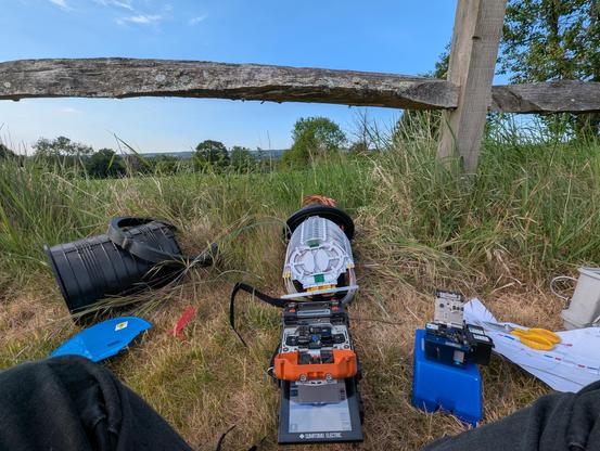A photograph taken by someone sitting on the ground, in front of them is a fibre splicing machine with an orange lid. Behind that device is the fibre enclosure with many trays of fibre. To the right is a fibre cleaver and the splicing map. On the left is the lid of the fibre enclosure. Behind all of that is a wooden bar fence and far in the distance are trees and blue sky.