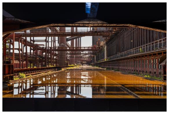 An industrial landscape featuring rusted metal structures and pipes, with a reflective water surface in the foreground. The scene suggests a deserted factory or manufacturing site, illuminated by sunlight.