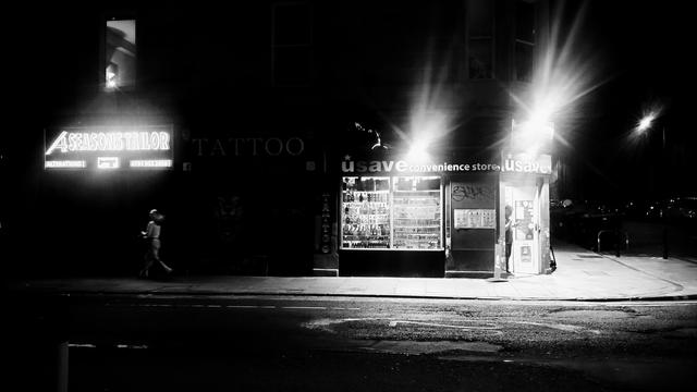 Black and white photo of a corner shop at night, with bright lights outside, contrasting with the darkness of the rest of the street