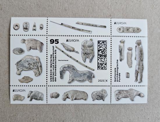 Special issue stamp from Germany featuring detailed images of Ice Age ivory figurines, flutes, and beads discovered in caves of the Swabian Jura. The stamp honors the UNESCO World Heritage site for prehistoric art and includes ancient animal carvings, human figures, and musical instruments. Text on the stamp reads “UNESCO-Welterbe Höhlen und Eiszeitkunst der Schwäbischen Alb” and “Nationale Archäologische Funde.” The stamp is marked "EUROPA," has a denomination of 95 Euro cents, and is dated 2025.